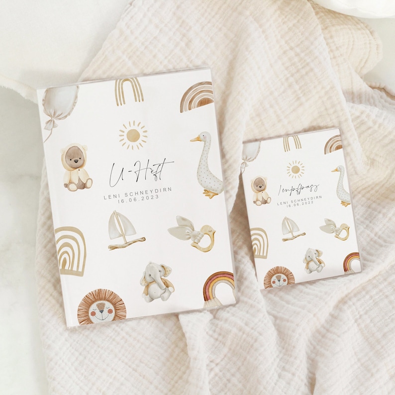 May include: Two white photo albums with a light brown border. The larger album has the text "U-H" followed by "LENI SCHNEYDERN 18.06.2023". The smaller album has the text "Lenipfags" followed by "LENI SCHNEYDERN 18.06.2023". Both albums have a white background with illustrations of a sun, a rainbow, a sailboat, a bird, a goose, a bear, an elephant, and a lion.