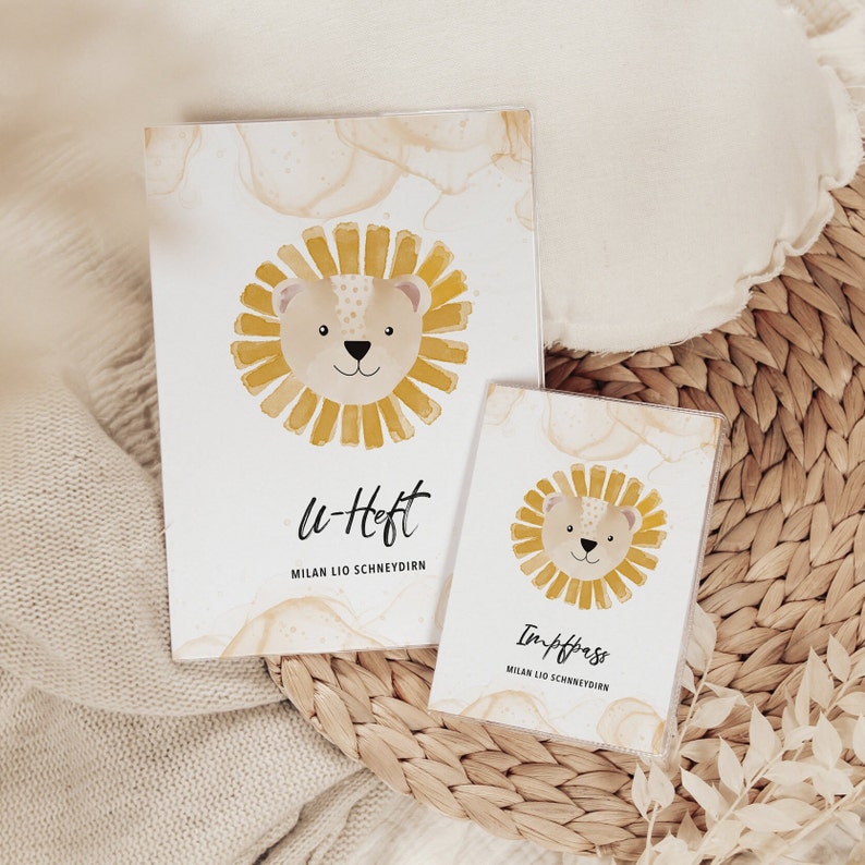 May include: Two white cards with a yellow lion illustration and text. The top card says "Lu-Heft" and "Milan Lio Schneydirn". The bottom card says "Impfpass" and "Milan Lio Schneydirn".