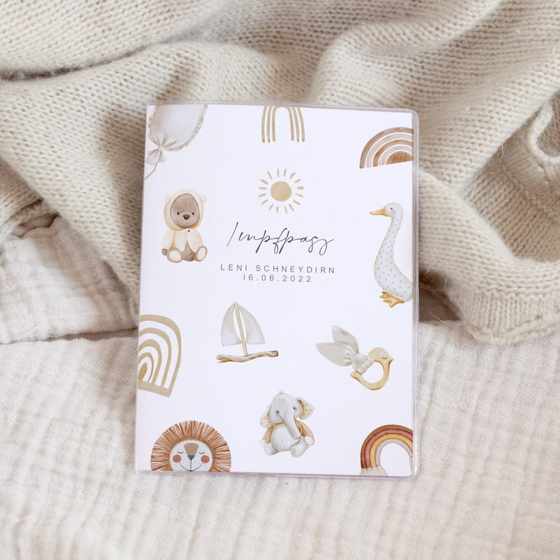 May include: A white card with a light brown and beige illustrated design. The card has the text "Impfpass" and "LENI SCHNEYDERN 16.06.2022". The illustrations include a sun, a rainbow, a teddy bear, a sailboat, a goose, a bird, an elephant, and a lion.