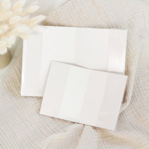 May include: Two white, rectangular, blank card covers with a subtle shimmer. The covers are lying on a white, textured fabric.