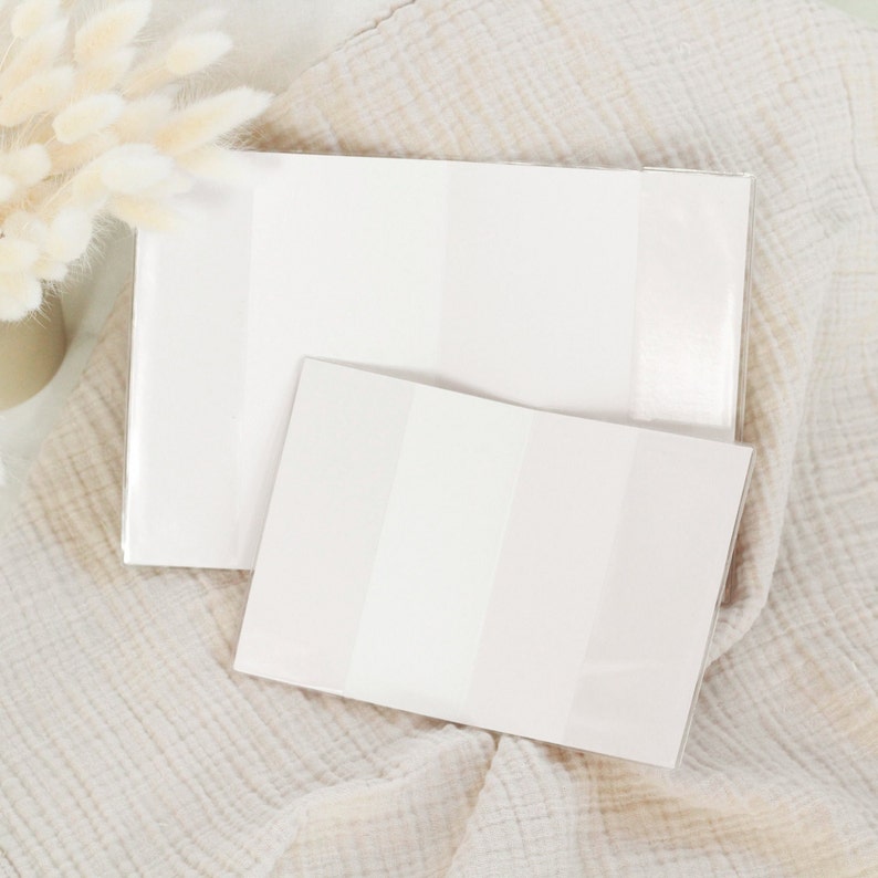 May include: Two white, rectangular, blank card covers with a subtle shimmer. The covers are lying on a white, textured fabric.