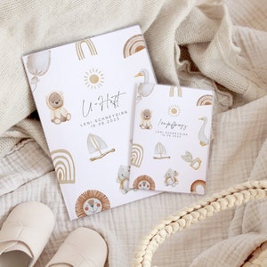 May include: Two white baby memory books with colorful illustrations of animals, rainbows, and suns. The larger book says "U-Heft" and "LENI SCHNEYDERN 18.06.2023". The smaller book says "Impfpass" and "LENI SCHNEYDERN 18.06.2022".