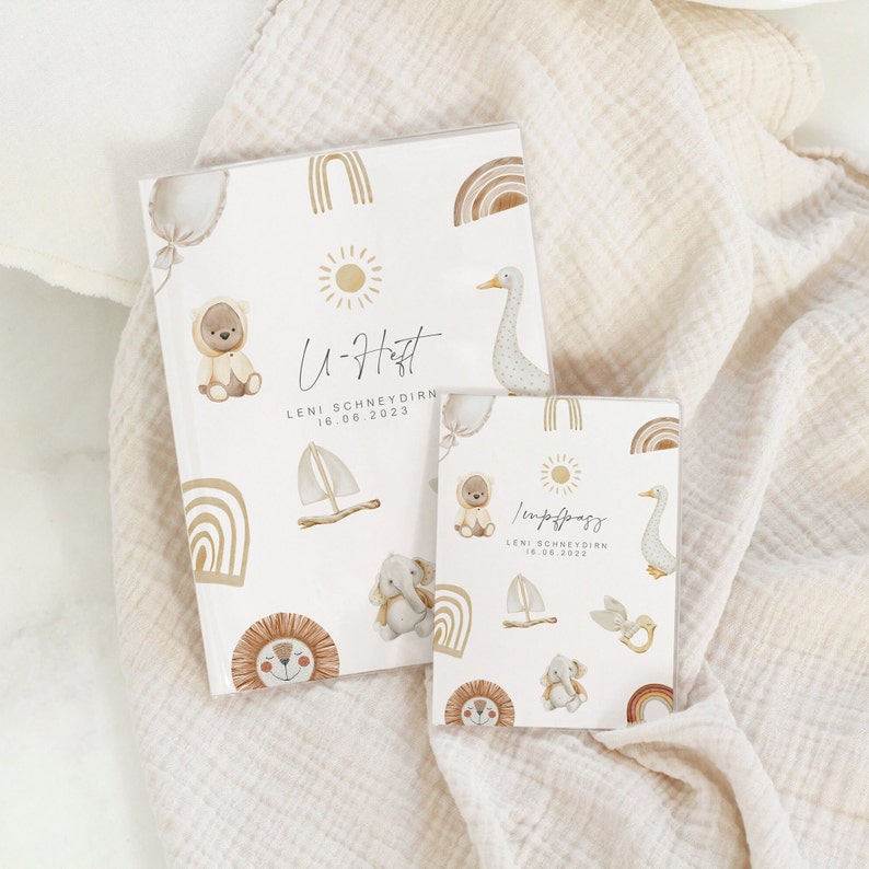 May include: Two white baby memory books with a light brown and gold design. The books have a sun, rainbow, sailboat, bear, lion, elephant, and goose illustrations. The text on the books reads "U-Haft Leni Schneyoirn 16.06.2023" and "Impfpass Leni Schneyoirn 18.08.2023".