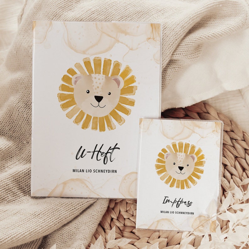 May include: Two white cards with a watercolor background featuring a cartoon lion with a yellow mane. The larger card says "u-Heft" and "MILAN LIO SCHNEYDIRN". The smaller card says "Impfpass" and "MILAN LIO SCHNEYDIRN".