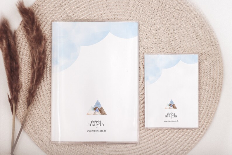 May include: Two clear acrylic frames, one large and one small, each containing a white print. The prints feature a blue watercolor cloud design at the top and a mountain illustration. The text "mein magda" and website address are at the bottom.