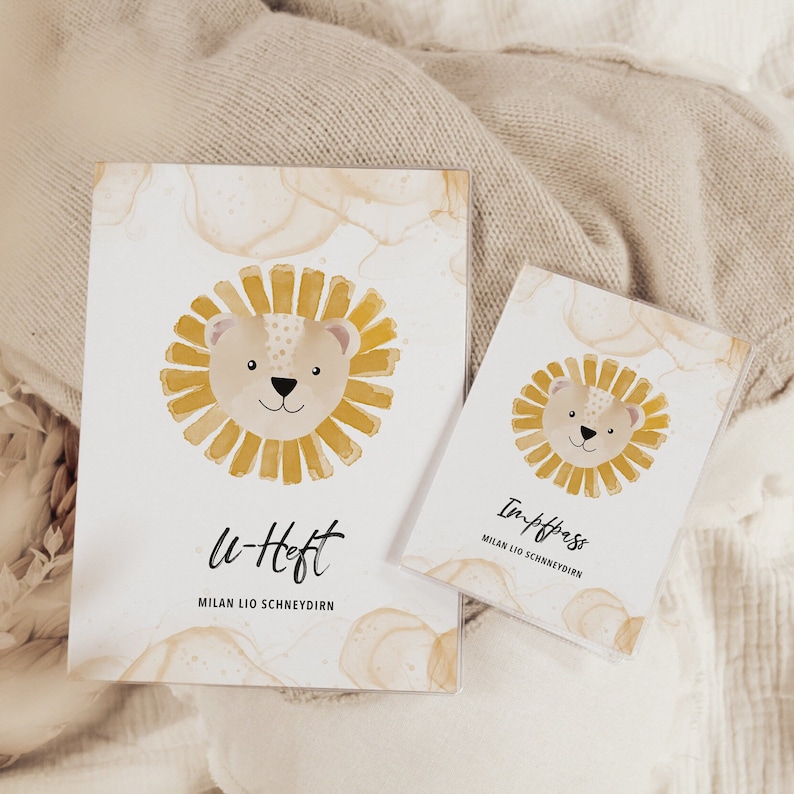May include: Two white notebooks with a watercolor background and a yellow lion illustration. The larger notebook says "u-Heft" and "Milan Lio Schneydirn". The smaller notebook says "Impfpass" and "Milan Lio Schneydirn".