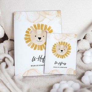 May include: Two watercolor lion illustrations on white cards with brown ink splatter. The larger card says "u-heft" and "MILAN LIO SCHNEY" below the lion. The smaller card says "Impfpass" and "MILAN LIO SCHNEYD" below the lion.