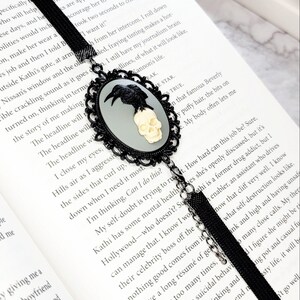 May include: Black bookmark with a silver skull and raven design. The bookmark is attached to a black ribbon with a tassel. The bookmark is resting on an open book with the text "I want it!" visible.