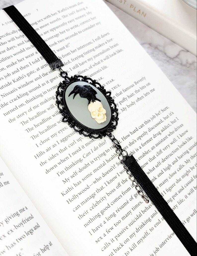 May include: Black bookmark with a silver skull and raven design. The bookmark is attached to a black ribbon with a tassel. The bookmark is resting on an open book with the text "I want it!" visible.