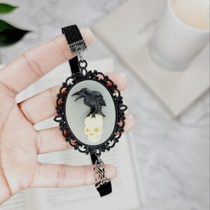 May include: Black crow perched on a white skull in a black ornate oval frame. The frame is attached to a black elastic bookmark.