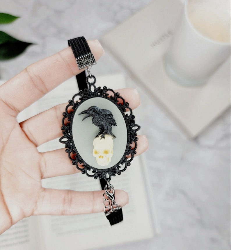 May include: Black crow perched on a white skull in a black ornate oval frame. The frame is attached to a black elastic bookmark.