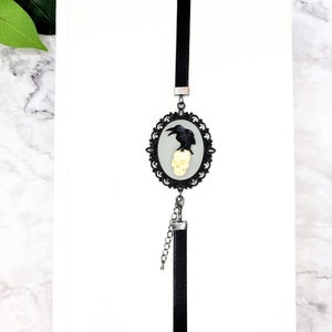May include: Black and white bookmark with a raven perched on a skull in a silver ornate frame. The bookmark has a black tassel.