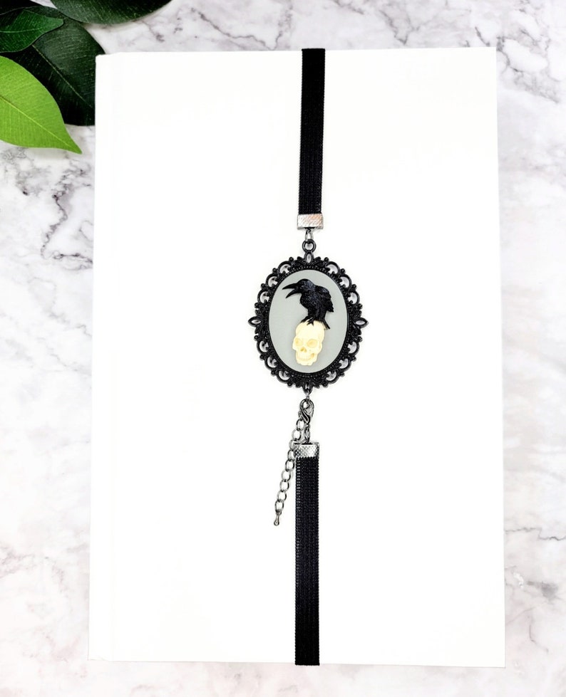 May include: Black and white bookmark with a raven perched on a skull in a silver ornate frame. The bookmark has a black tassel.