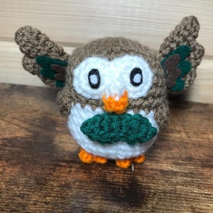 May include: A crocheted stuffed owl toy with brown and white fur, green wings, and orange feet. The owl is holding a green leaf in its beak.