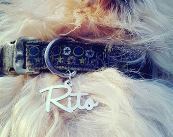 May include: A silver-toned metal pet tag shaped in a cursive script reading Rito, attached to a dark-colored fabric collar featuring a pattern of small blue and green stars and circles, worn around the neck of a long-haired, light-colored animal.