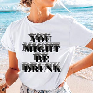 May include: White t-shirt with the text "YOU MIGHT BE DRUNK" in a layered black and gray font. The shirt is short-sleeved and worn by a person on a beach with the ocean in the background.