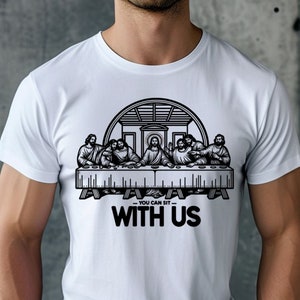 May include: White t-shirt featuring a black and white graphic of the Last Supper. The design includes the text "YOU CAN SIT WITH US" below the image. The shirt is made of a soft material.