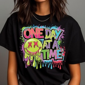 Könnte beinhalten: Schwarzes T-Shirt mit einem farbenfrohen Grafikdesign. Das Design zeigt den Text "ONE DAY AT A TIME" in einer lebendigen, mehrfarbigen Schriftart, zusammen mit einem grünen Smiley mit tropfendem Schleim und roten "XX"-Augen.