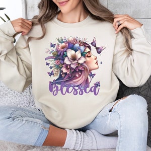 Op de afbeelding: Een crèmekleurige trui met een paars bloemenontwerp en een vrouwengezicht. Het ontwerp bevat vlinders en het woord "blessed" in paars.