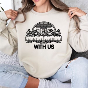 May include: Cream-colored sweatshirt featuring a black graphic of the Last Supper. The graphic includes the text "YOU CAN SIT WITH US" in a bold, black font. The sweatshirt is a casual, long-sleeved garment.