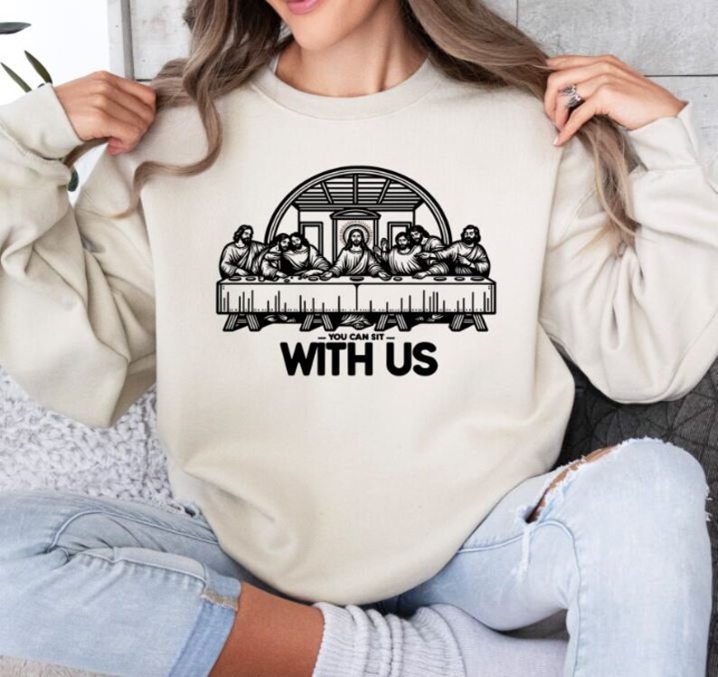 May include: Cream-colored sweatshirt featuring a black graphic of the Last Supper. The graphic includes the text "YOU CAN SIT WITH US" in a bold, black font. The sweatshirt is a casual, long-sleeved garment.