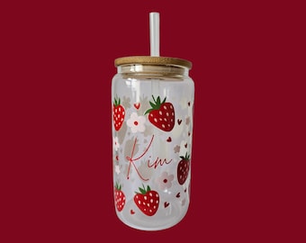 May include: A clear glass tumbler with a bamboo lid and a white straw. The tumbler is decorated with red strawberries, white flowers, and the name "Kim" in a cursive font. The background is a solid dark red.