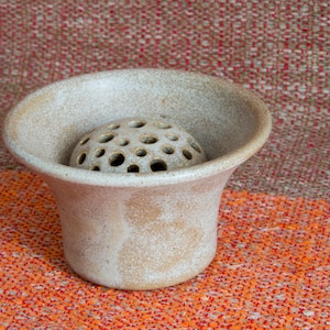 Könnte beinhalten: Eine kleine, weiße Keramikvase mit einem runden, perforierten Boden. Die Vase hat eine gesprenkelte, strukturierte Oberfläche und steht auf einem roten und orangefarbenen Stoff.