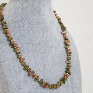 Puede incluir: Un collar hecho de cuentas de piedra verde, roja y marrón. Las cuentas son de forma y tamaño irregulares, y están enhebradas en una cadena dorada.