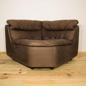 Op de afbeelding: Een bruine leren hoekbank met een afgeronde vorm. De bank heeft een laag profiel en een ontwerp met twee kussens. Het leer lijkt versleten, wat het een vintage uitstraling geeft. De bank staat op een houten vloer.