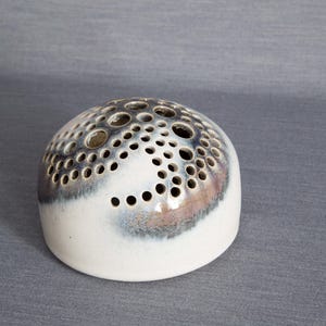 Op de afbeelding: Witte keramische bloempot met een gespikkelde glazuur en een patroon van kleine gaatjes. De pot is rond en heeft een gewelfde bovenkant.