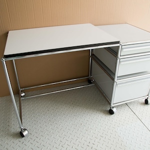 Könnte beinhalten: Moderner Schreibtisch mit weißer Tischplatte und silberfarbenem Metallrahmen. Er hat drei Schubladen auf der rechten Seite und ist auf Rollen montiert. Der Schreibtisch ist für ein Homeoffice oder einen Arbeitsbereich konzipiert, ca. 120 cm lang.