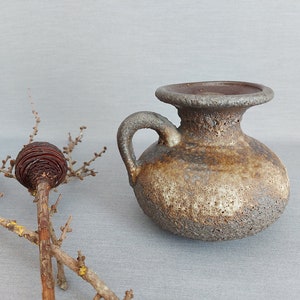 Puede incluir: Un pequeño jarrón redondo de cerámica con asa y borde ancho y plano. El jarrón tiene una superficie texturizada y moteada en tonos marrón, beige y gris. Un tallo de planta seca con una piña está en primer plano.
