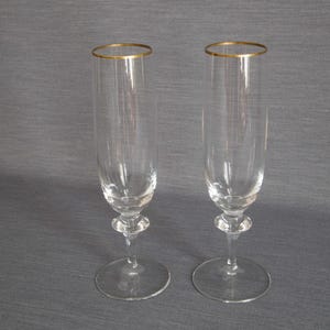 May include: Two clear glass champagne flutes with gold rims. The flutes are on a grey background.