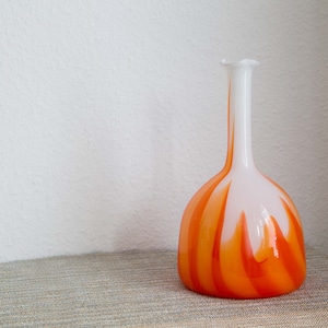 Könnte beinhalten: Eine weiße und orangefarbene Glasvase mit einer ausgestellten Öffnung und einem abgerundeten Boden. Die Vase hat ein wirbelndes Muster aus Orange und Weiß.