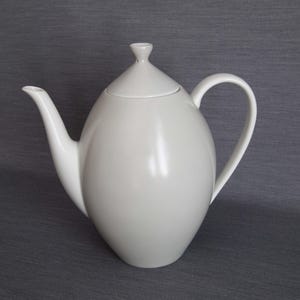 May include: A white ceramic teapot with a lid and a curved handle. The teapot has a rounded body and a spout. The lid has a small knob on top. The teapot is on a grey background.