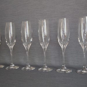 May include: Six clear glass champagne flutes with a delicate, fluted design. The flutes are arranged in a row on a grey surface.