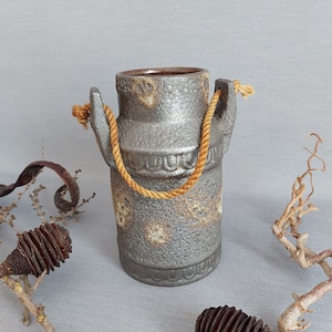 Puede incluir: Un jarrón de cerámica gris con un asa de cuerda, decorado con un patrón floral. El jarrón está sobre una superficie gris.