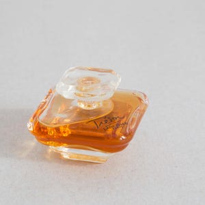 Könnte beinhalten: Eine durchsichtige Glasflasche mit goldfarbenem Parfüm. Die Flasche hat einen dekorativen Verschluss und der Text "Tresor" ist auf der Vorderseite aufgedruckt.