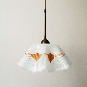 Puede incluir: Una lámpara colgante blanca con una pantalla plisada y un vástago de color bronce. La pantalla tiene un borde festoneado con un diseño geométrico en color cobre. La luz cuelga del techo.