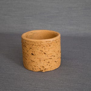 Könnte beinhalten: Ein kleiner, zylindrischer Terrakotta-Pflanztopf mit einer rauen, strukturierten Oberfläche. Der Topf ist hellbraun und hat einen leicht unebenen Rand.