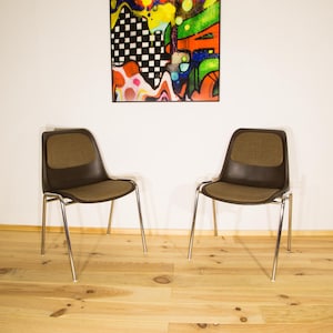Può includere: Due sedie marroni e beige con gambe cromate sono posizionate su un pavimento in legno chiaro. Sopra, un colorato dipinto astratto è appeso a una parete bianca. Le sedie hanno un design moderno della metà del secolo.