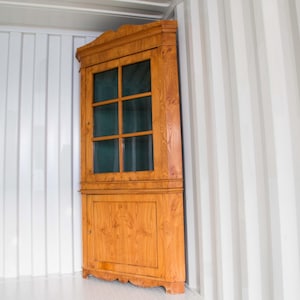 Puede incluir: Un mueble de esquina de madera con un acabado marrón claro. El mueble tiene una sección superior con paneles de vidrio con seis paneles y una sección inferior sólida. El mueble está en una habitación blanca.