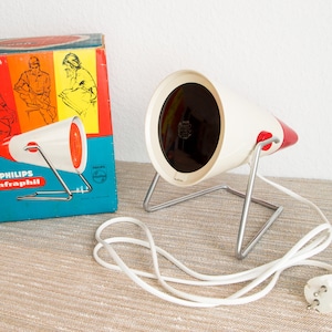 Puede incluir: Una lámpara de calor infrarroja Philips Infraphil blanca y roja con un reflector negro. La lámpara está sobre un soporte metálico y tiene un cable blanco con un enchufe europeo.