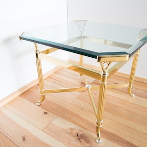 Puede incluir: Una mesa con una tapa de vidrio transparente y una base de metal dorado. La mesa tiene una tapa de vidrio transparente y una base de metal dorado con un diseño geométrico.