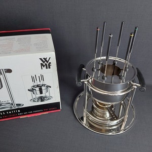 Può includere: Un set di fonduta in argento con una pentola rotonda e otto forchette per fonduta. Il set è su un supporto con una base. La scatola del set è sullo sfondo e dice "Fondue 11 teilig" e "WMF" su di essa.