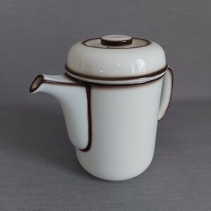 Op de afbeelding: Witte theepot van keramiek met een bruine rand en handvat. De theepot heeft een deksel met een bruine knop.