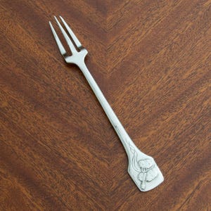 以下が含まれることがあります： 木製の表面に置かれた、3つの歯が付いた銀色のフォーク。ハンドルには、花の要素を持つ人物のエンボス加工のデザインがあります。フォークは銀食器の一部です。