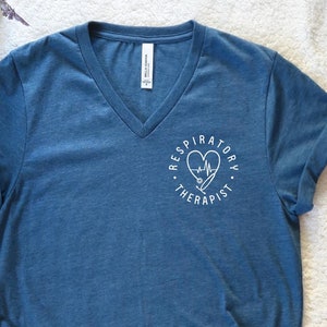May include: A blue V-neck t-shirt with rolled-up sleeves. The shirt features a white graphic on the chest that says "Respiratory Therapist" around a heart, stethoscope, and heartbeat line. The shirt is knotted at the bottom.