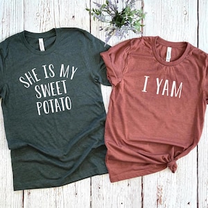 May include: Two short-sleeved t-shirts. One is dark gray with the text "SHE IS MY SWEET POTATO" in white. The other is a reddish-brown color with the text "I YAM" in white. Both shirts are on a light wood background.