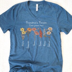 May include: Blue V-neck t-shirt with the text "Grandma's Garden Love grows here" and floral illustrations. The shirt features names like Archie, Deborah, and Joshua beneath the flowers. A casual, comfortable tee.